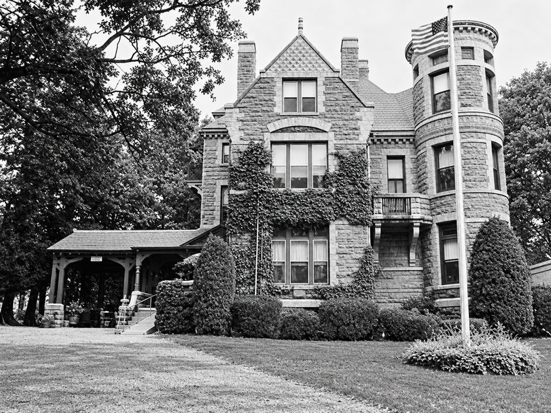 Black-and-white photograph of the stone house behind a lawn and flagpole, ivy on the front, the wooden porte-cochère at left