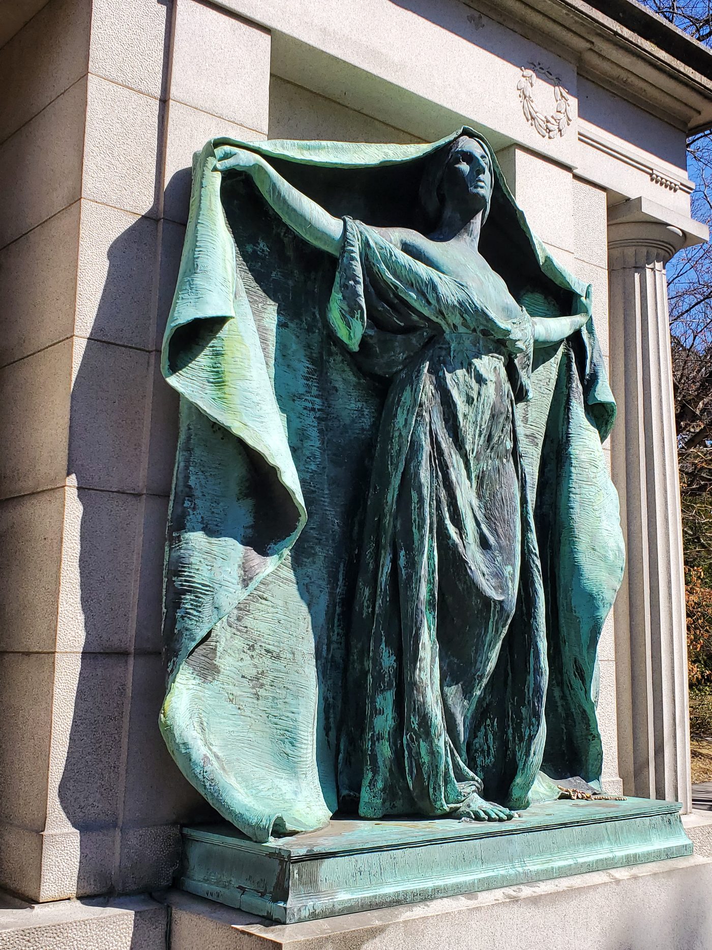 A green-patinated bronze figure of a woman with raised arms holding a long drape, set in a pale granite surround, in daylight