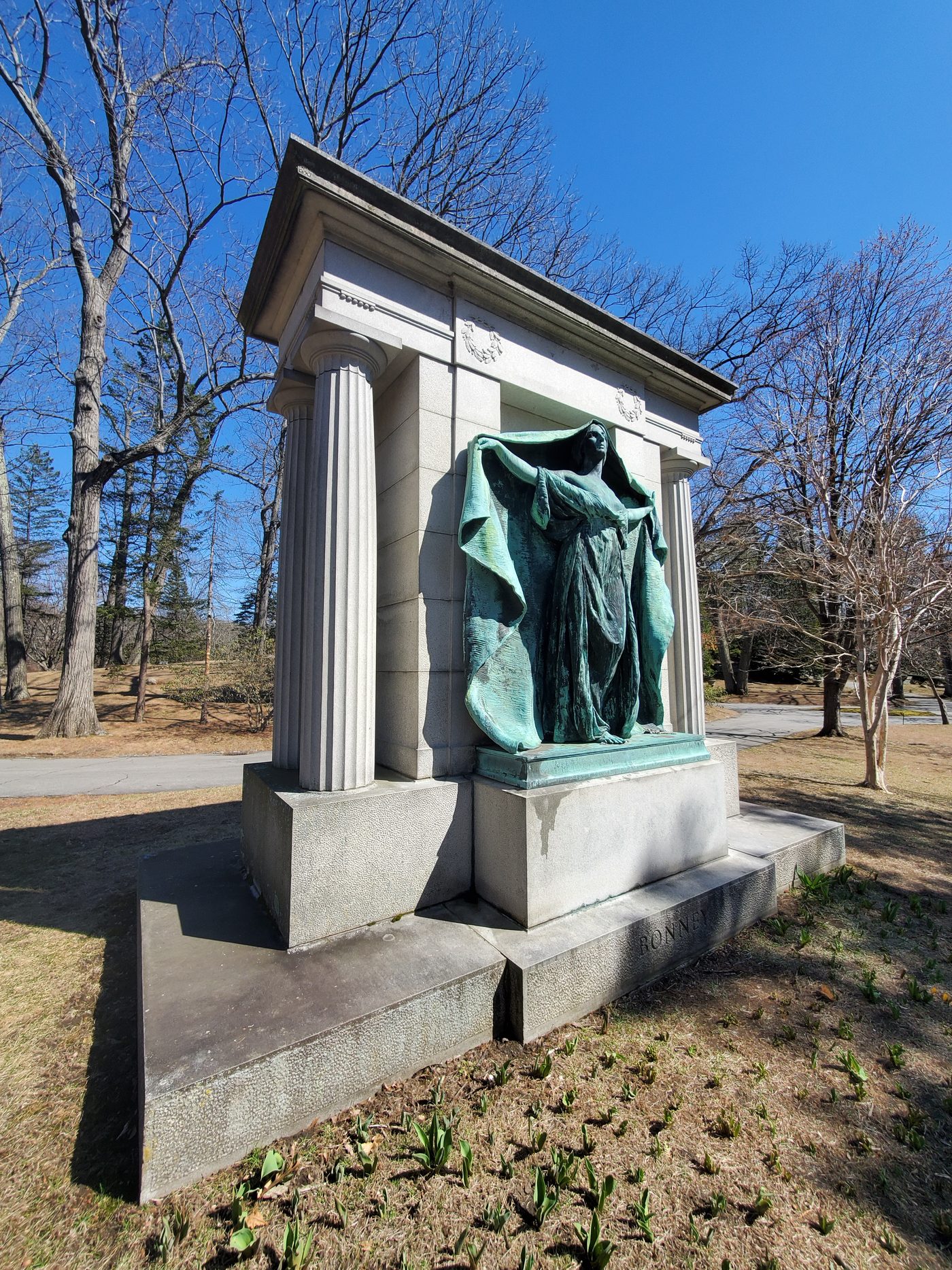 The whole monument from the front: a granite surround with columns framing the bronze figure, bare trees and blue sky behind