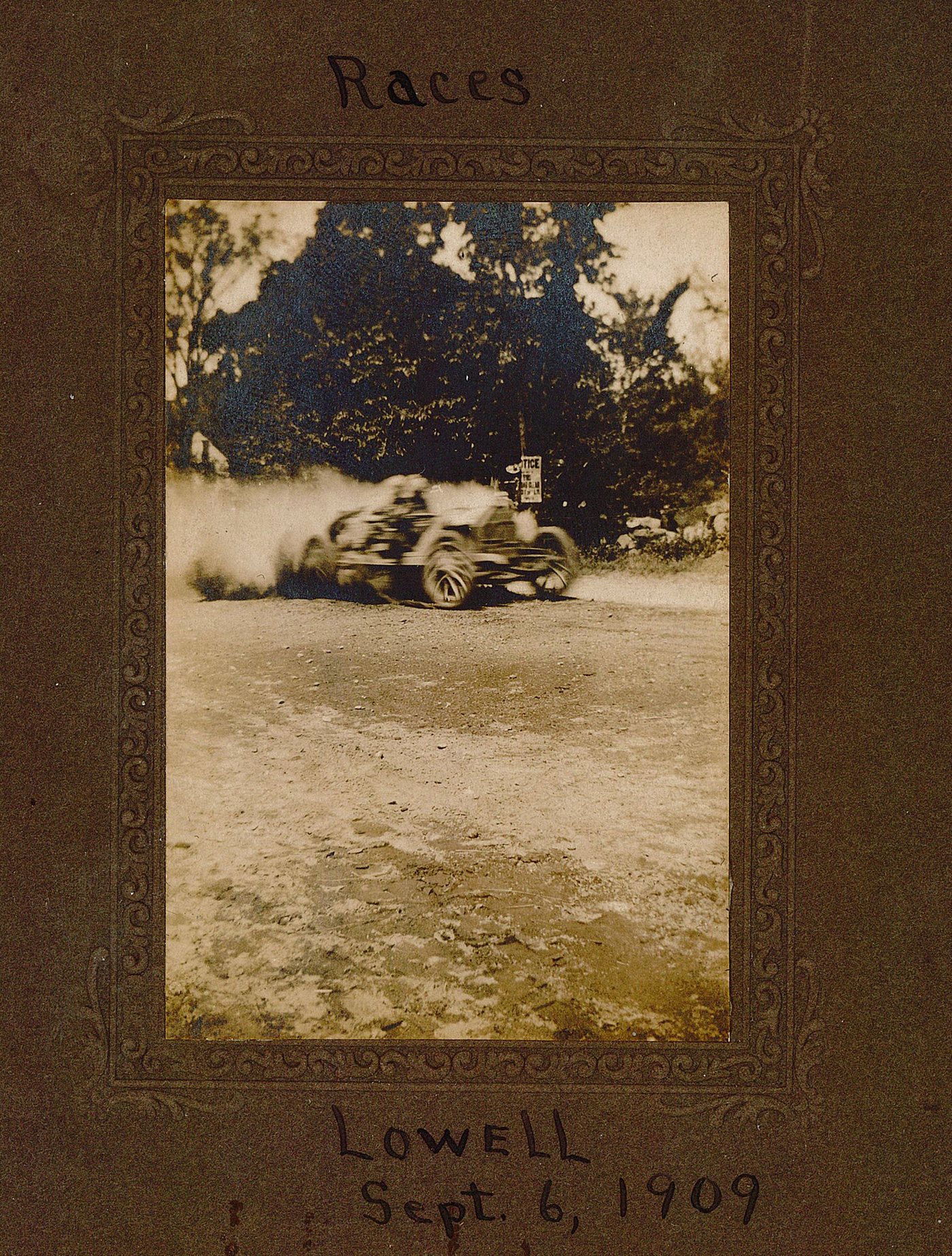 A racing car raising dust on a dirt road, in a photograph mounted on a card inscribed “Races, Lowell, Sept. 6, 1909”