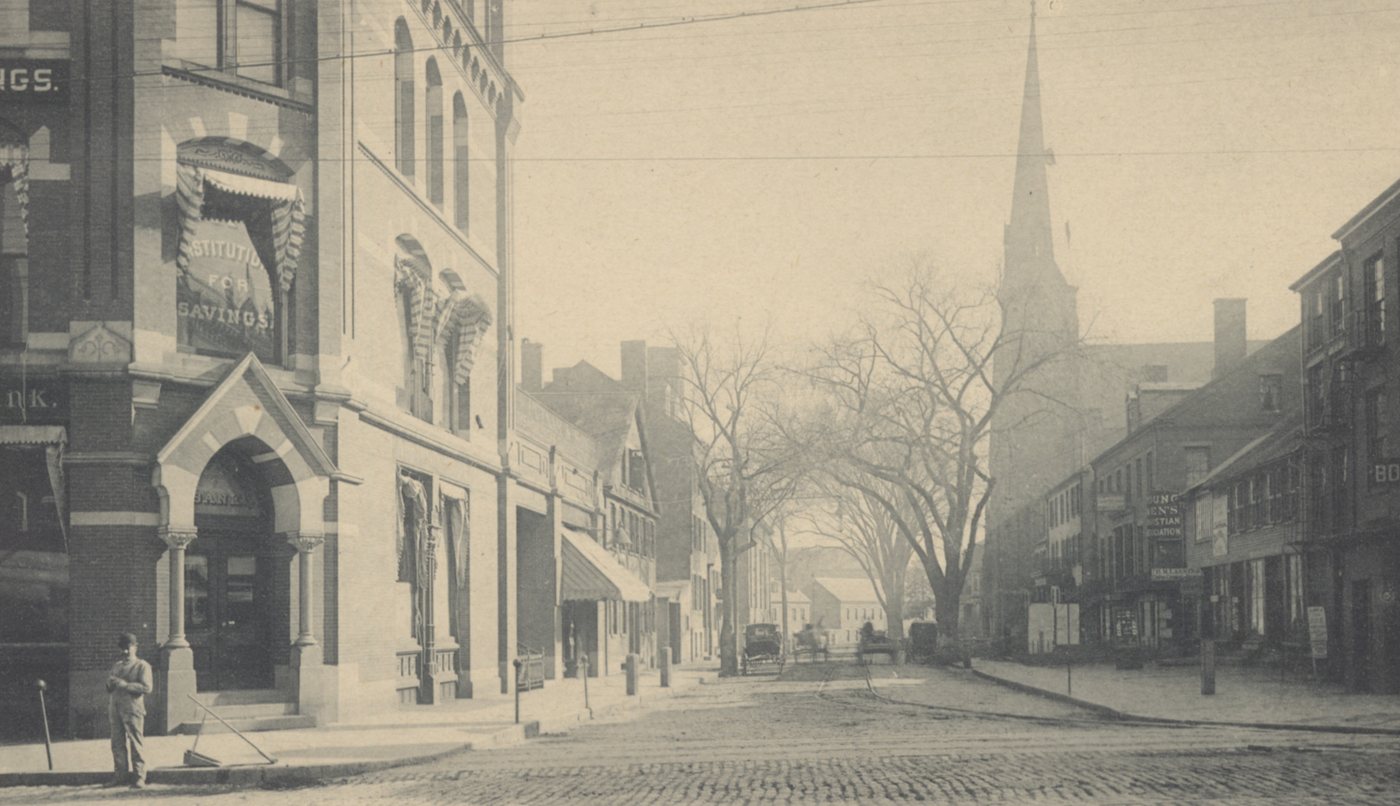 A street corner with an arched bank entrance, a cobbled street and a church spire in the distance
