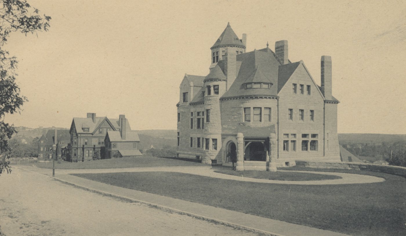 Large towered stone house on a lawn at a curving street corner, with other houses beyond