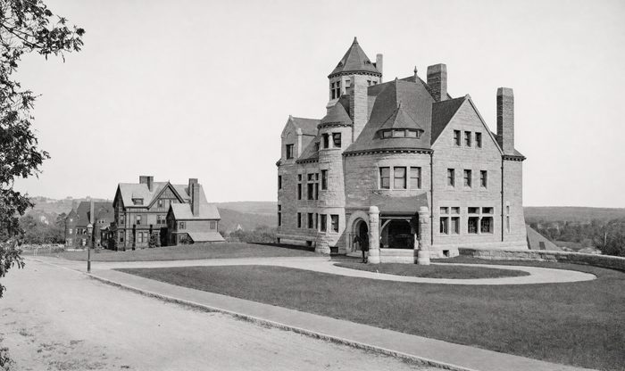 Large towered stone house on a lawn at a curving street corner, with other houses beyond