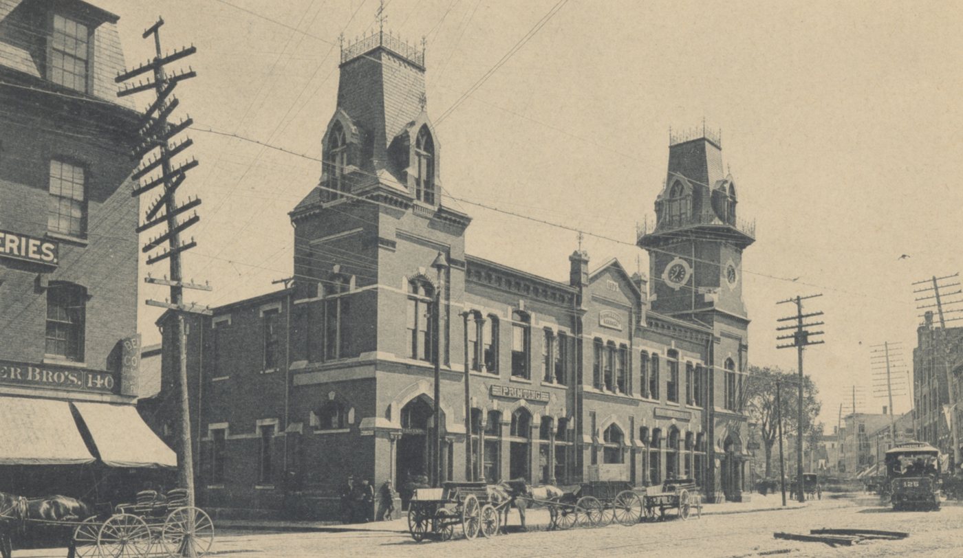 A railroad station with two towers, with carriages and wagons lined up in the street