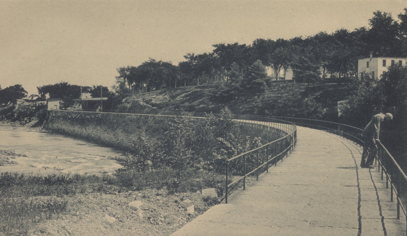 A railed walkway curving along the bank of a canal beside the river, with a figure at the rail