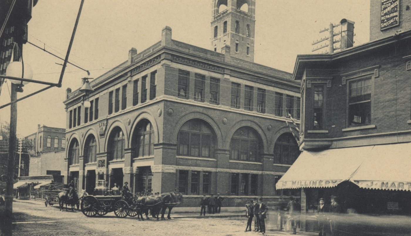A brick fire station with arched doors and a tower, with horse-drawn wagons in the street