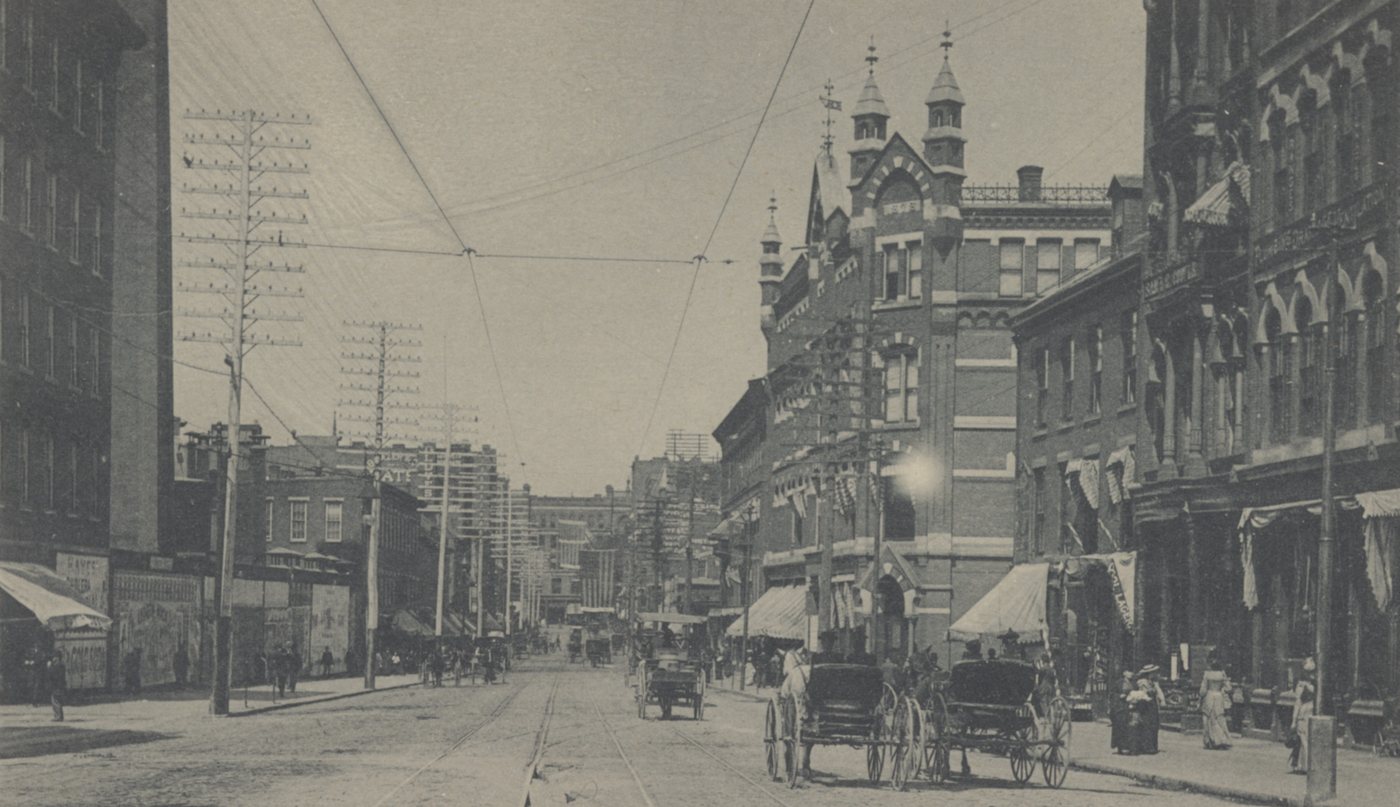 A street of tall brick commercial buildings with awnings, wagons and overhead wires