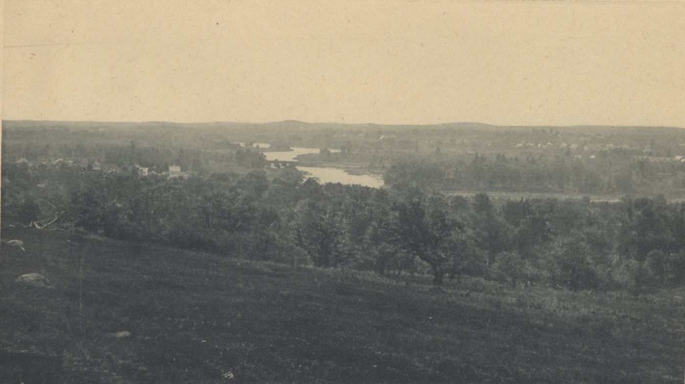 Wide view from a grassy hillside over trees to the winding Concord River and distant hills