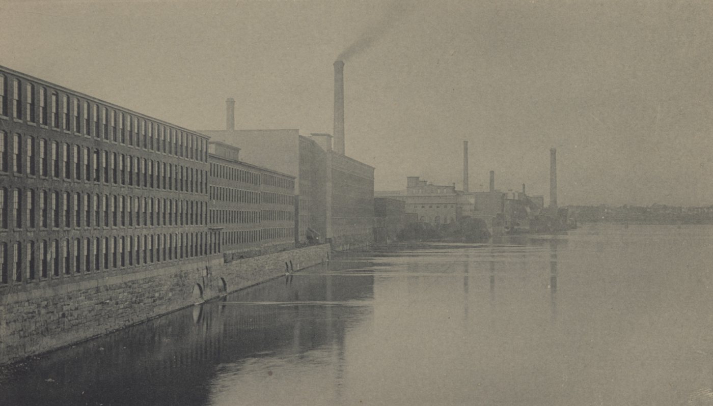 A long row of mill buildings and smokestacks along the river, reflected in still water