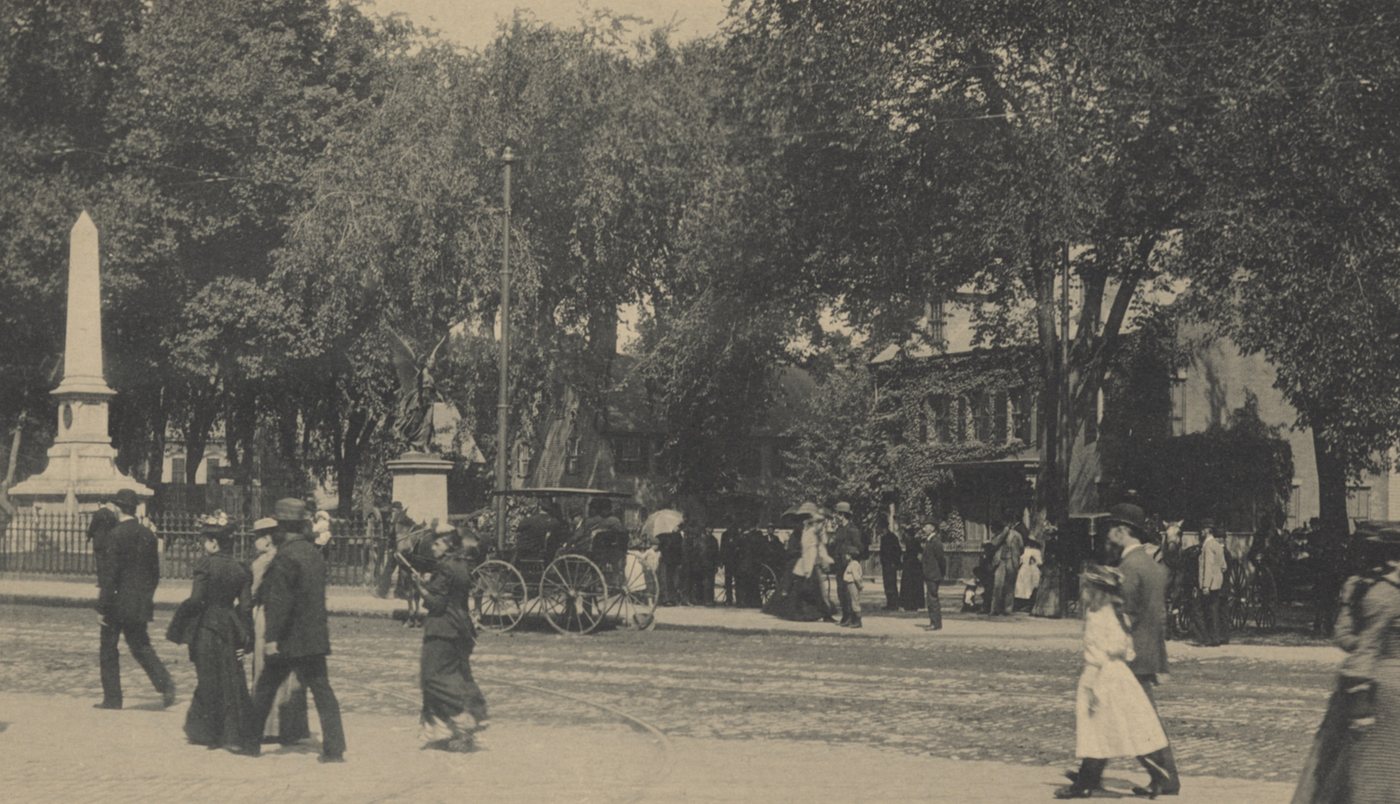 People walking across a square in front of an obelisk and trees, with a horse and carriage at the curb