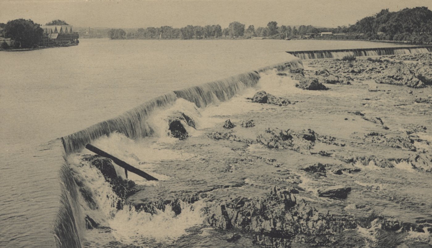 Water pouring over a dam and down a broad ledge of rock, with the river beyond