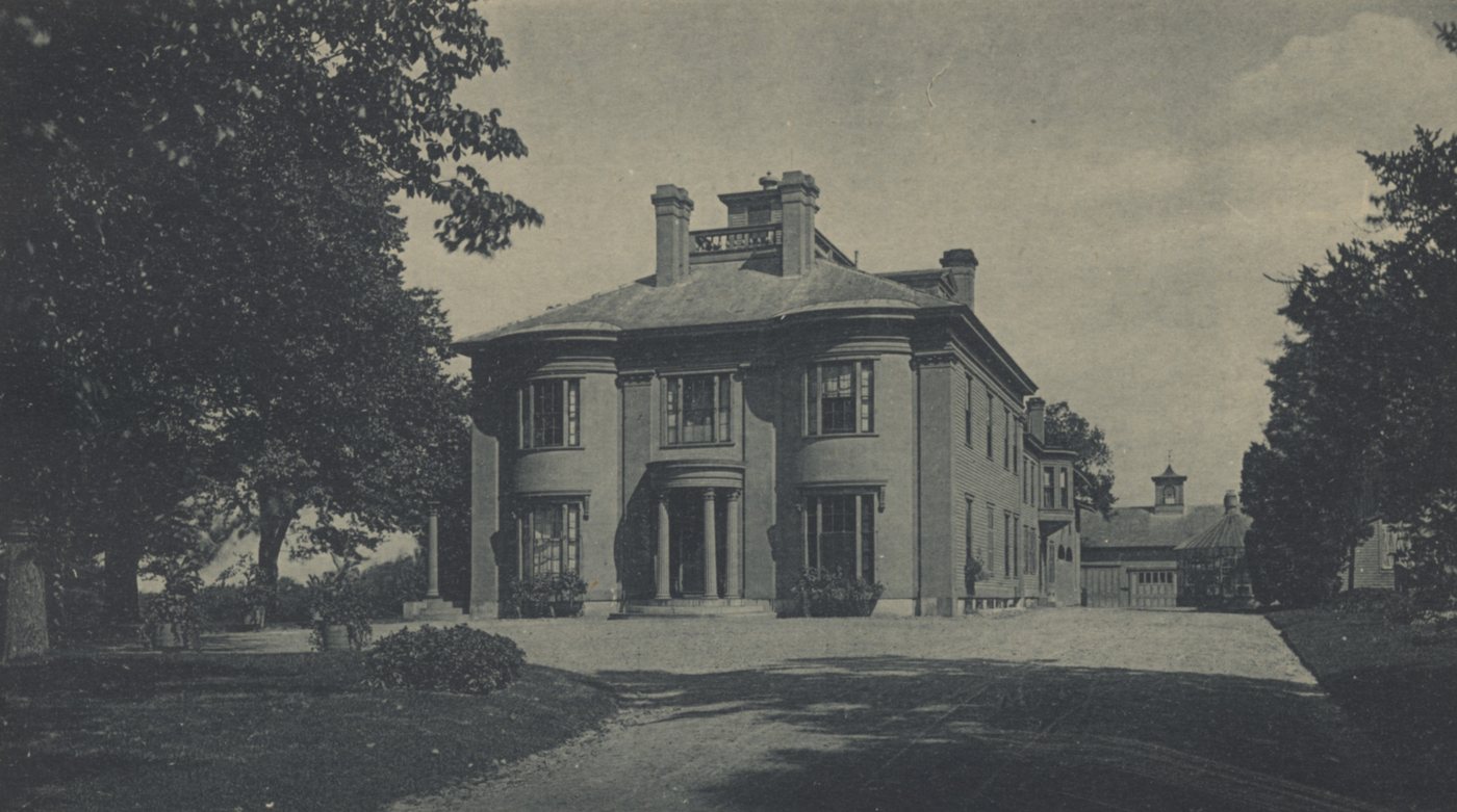 A square stone mansion behind a lawn and drive, framed by large trees