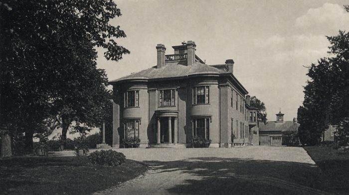 A square stone mansion behind a lawn and drive, framed by large trees