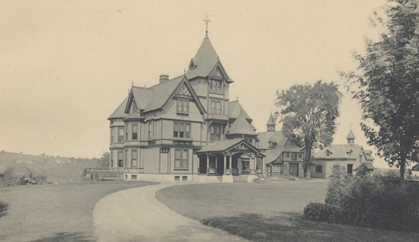 A large shingled house with a tower and steep gables at the end of a curving drive