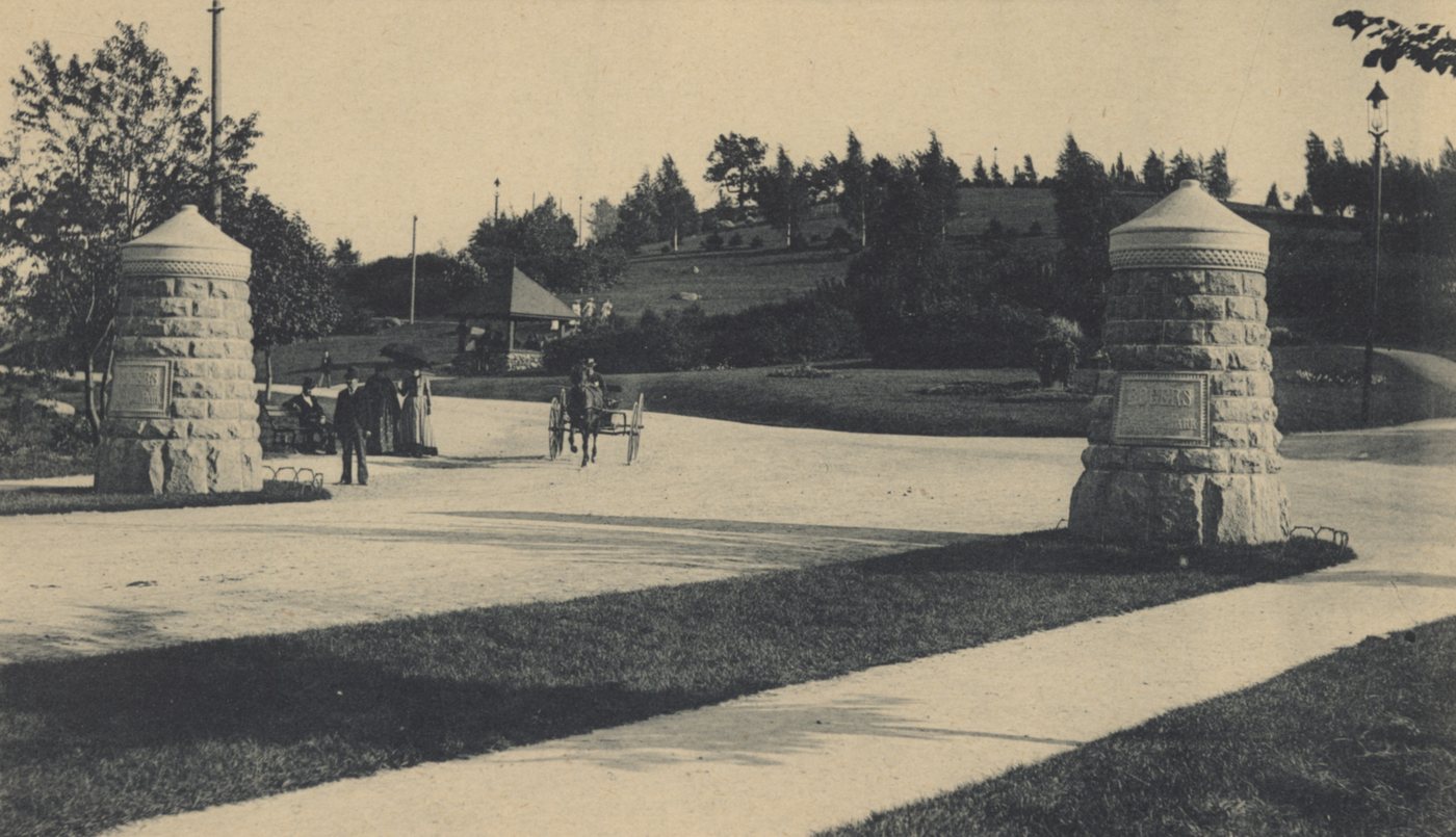 Two stone gate towers at the entrance of a park, with a horse and carriage on the road beyond