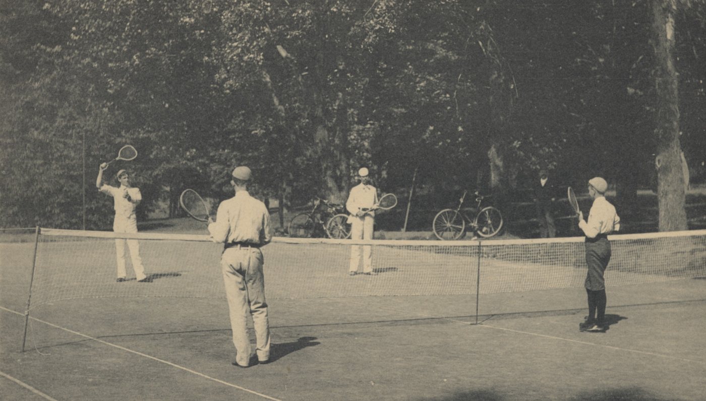 Four men in white playing tennis on a grass court under tall trees, bicycles at the back