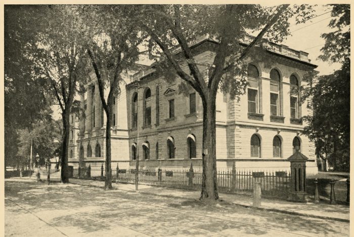 A classical stone courthouse behind tall trees and an iron fence