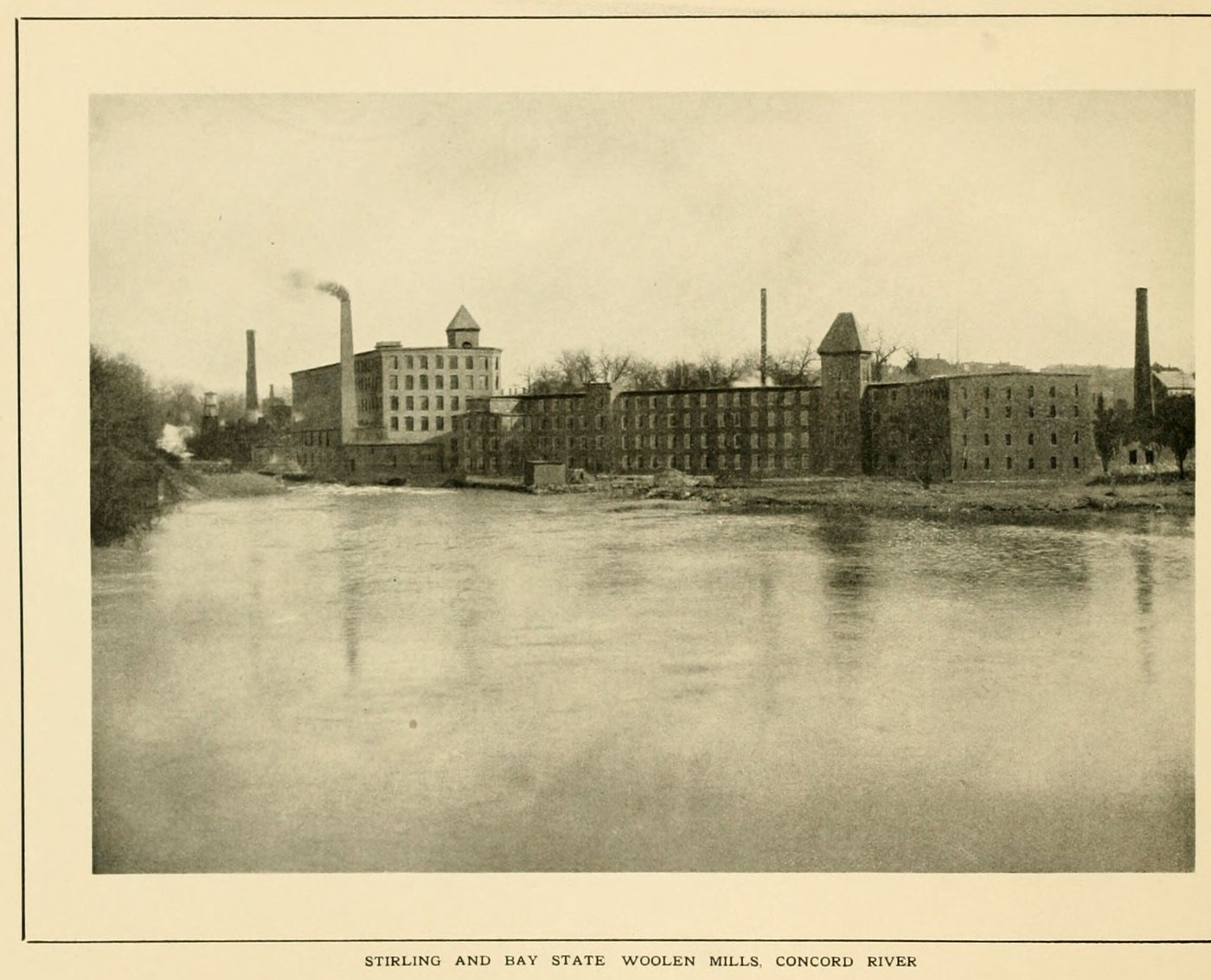 Brick mill buildings with a tower and chimneys across a wide stretch of river
