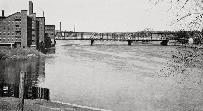 A steel truss bridge across the river with brick mill buildings on the far bank