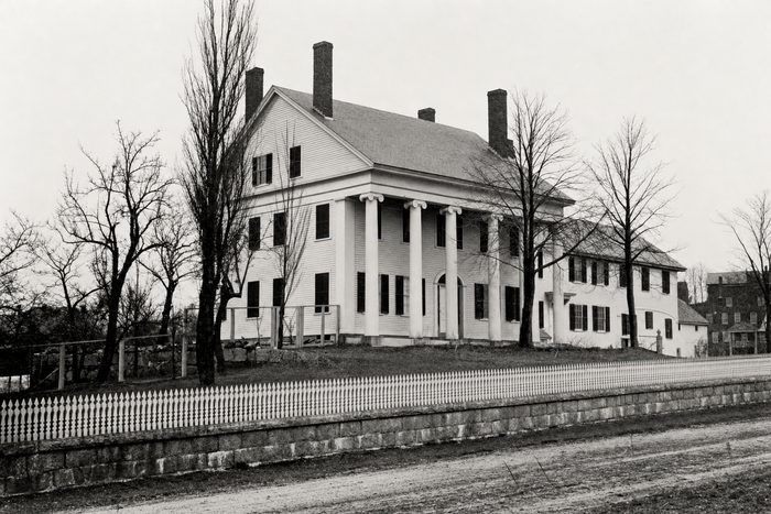 A white columned house with two chimneys behind a fence and bare trees