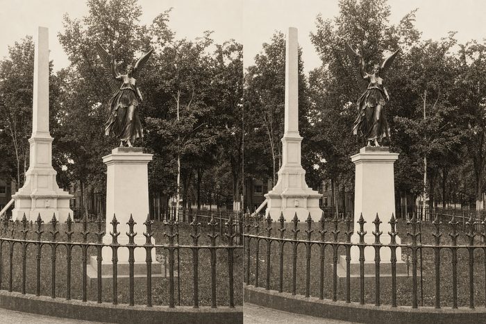 Stereograph of a tall obelisk and a statue on a pedestal behind an iron fence, with trees
