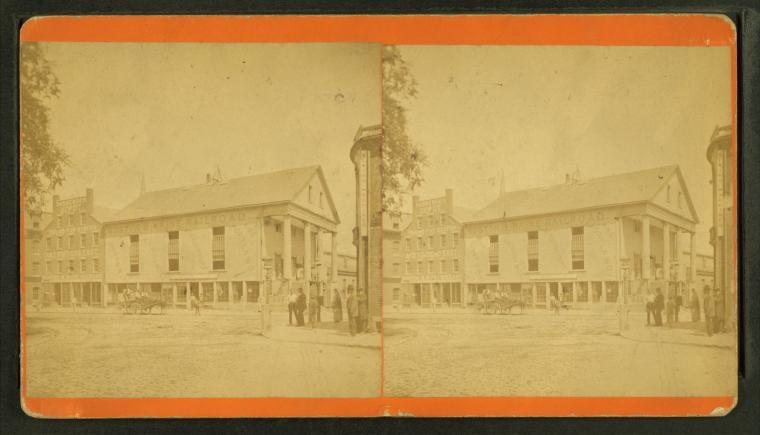 Stereograph of a street corner with a columned building and a row of storefronts