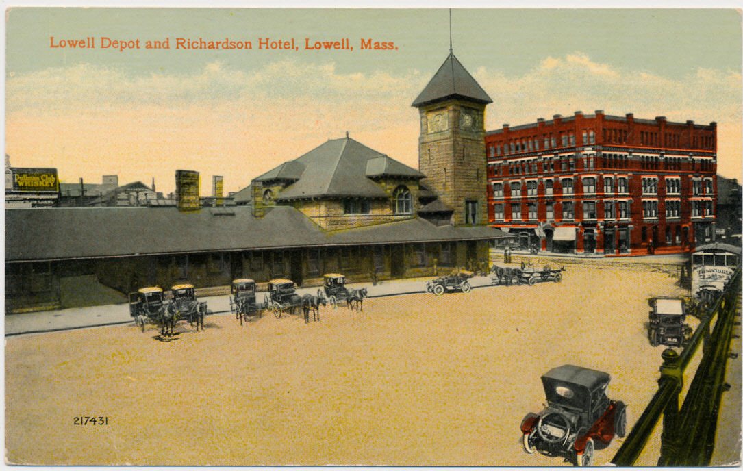 A color postcard of a depot with a clock tower and a brick hotel, with carriages and an open automobile in the foreground