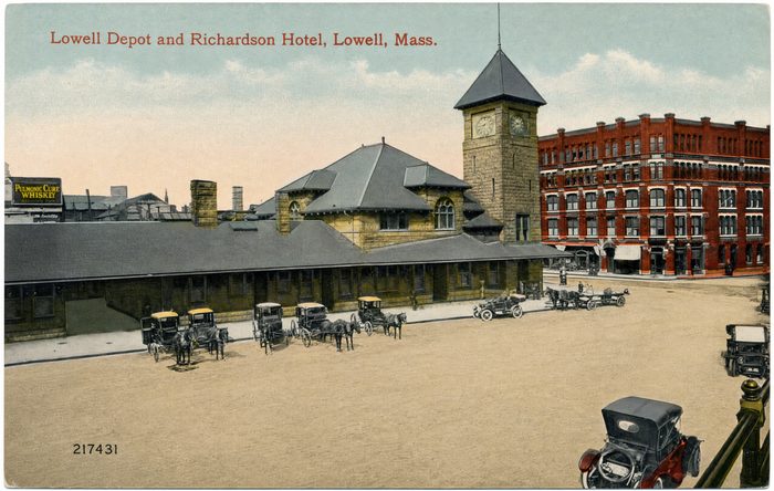 A color postcard of a depot with a clock tower and a brick hotel, with carriages and an open automobile in the foreground
