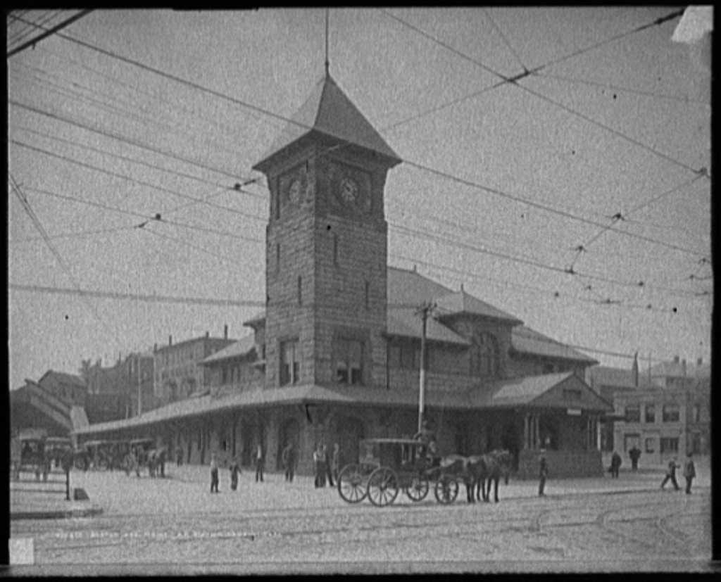 A railroad station with a tall clock tower, horse-drawn carriages waiting and streetcar wires overhead