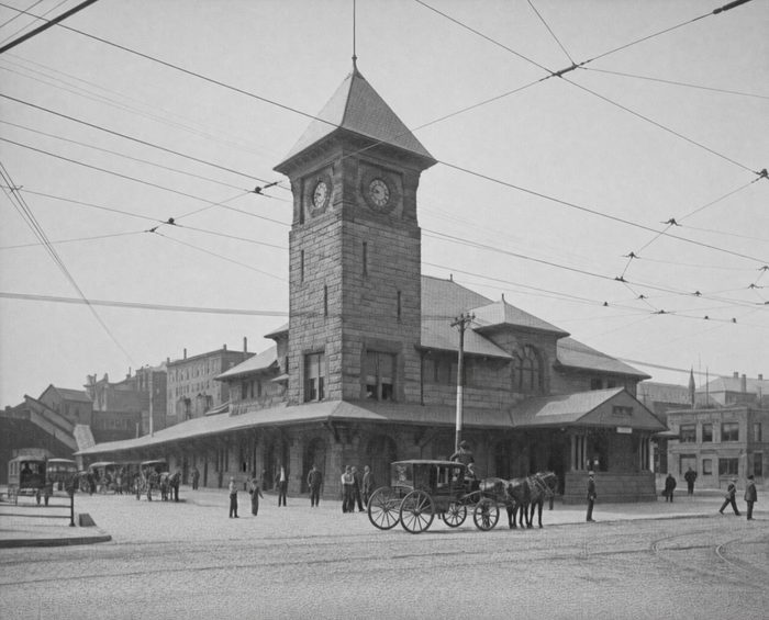 A railroad station with a tall clock tower, horse-drawn carriages waiting and streetcar wires overhead
