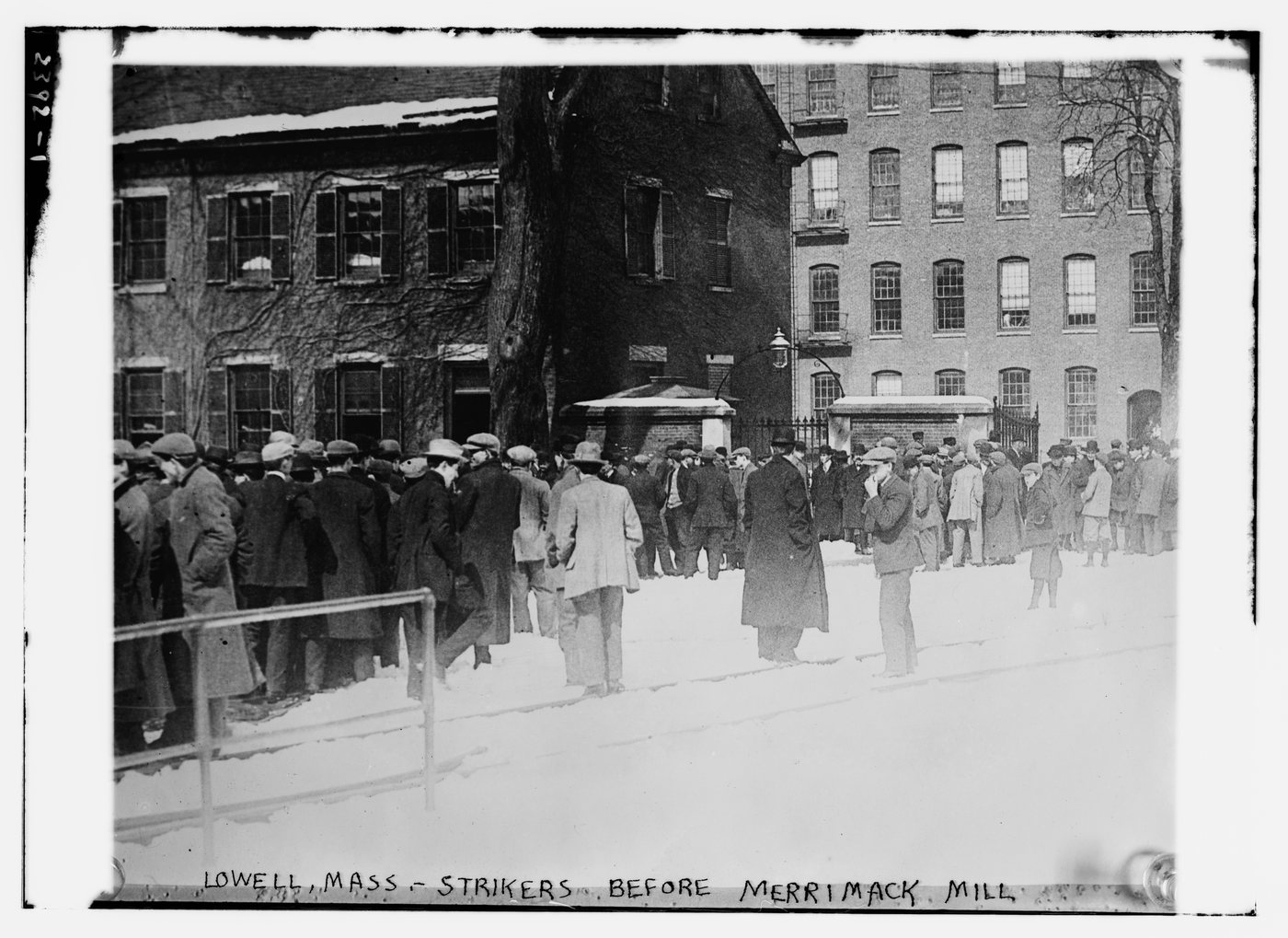 A crowd of men in hats and overcoats standing in the snow in front of a brick mill
