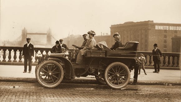 A man and a woman seated in an early open motor car on a London street