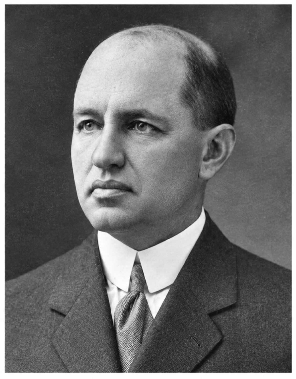 Black-and-white portrait of a bald man in middle age with a broad face, in a dark suit, white collar and patterned tie, three-quarter view