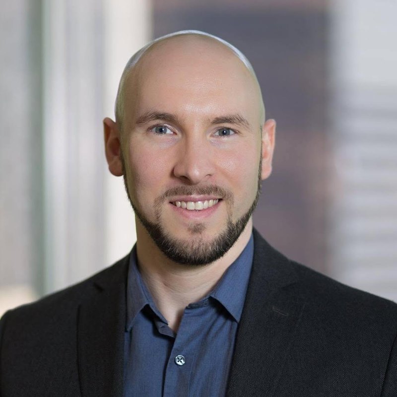 Head-and-shoulders photograph of a smiling bald man with a short beard, in a dark jacket and blue shirt