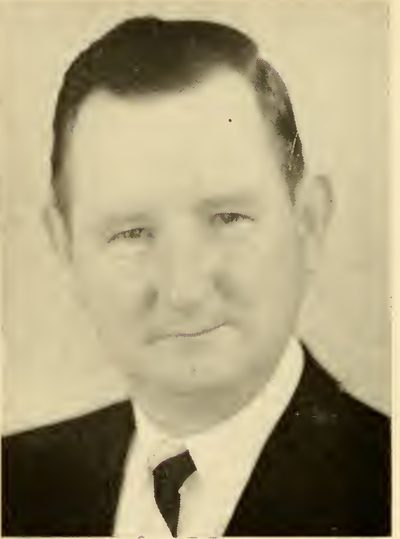 Halftone photograph of a man in his late forties with dark, side-parted hair, in a dark suit and tie, facing the camera