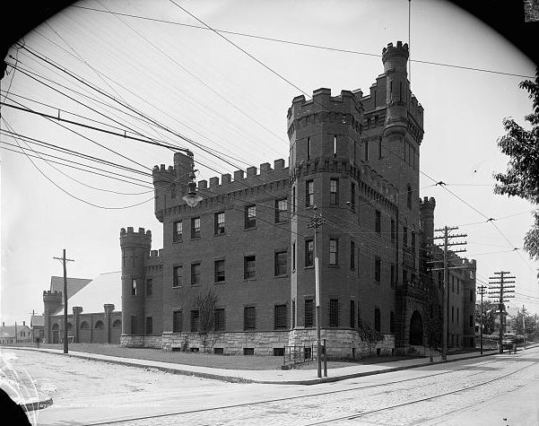 The State Armory in Lowell about 1908: a brick fortress on a stone base with crenellated round towers and a tall corner turret, overhead wires and a streetcar track in front