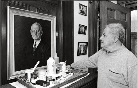 Black-and-white photograph of a gray-haired man in a sweater in a wood-paneled room, looking up at a framed portrait of an older man in a suit